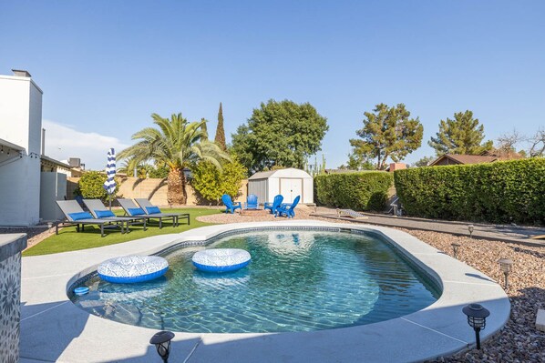 Une piscine dotée de chaises longues et d’un jacuzzi, entourée d’un jardin aménagé avec des arbres et d’un bâtiment blanc.