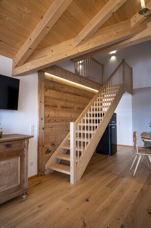 A wooden staircase leading to an upper level, a flat-screen TV mounted on the wall, and a wooden cabinet with drawers. - Ferienwohnung Singer "morgensonne" (Wackersberg)