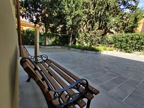 Um banco de madeira com apoio de braço de metal numa área externa pavimentada, com árvores e um edifício ao fundo.