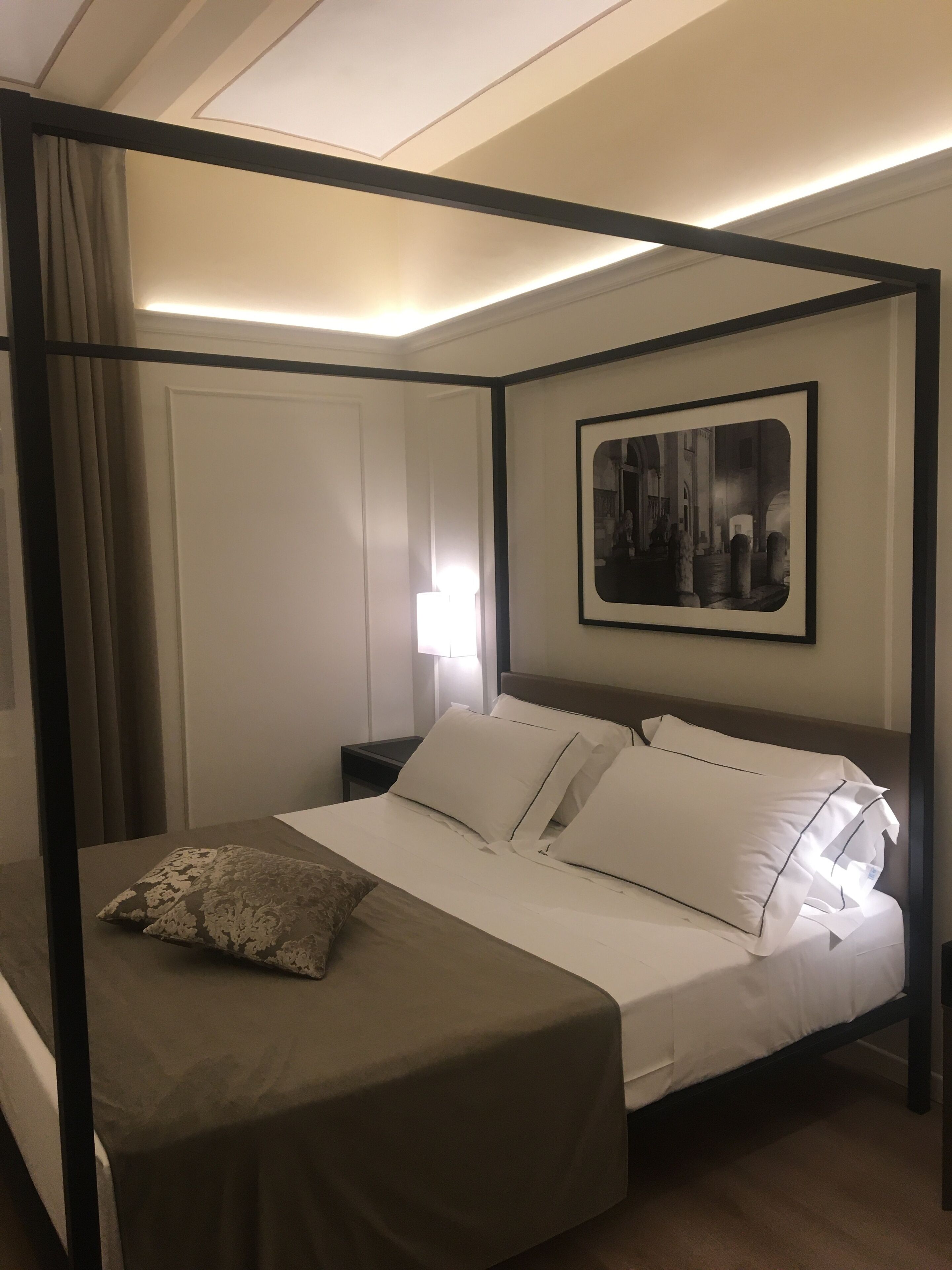 A four-poster bed with white linens and a single decorative pillow. A framed black and white photograph is on the wall above the bed.