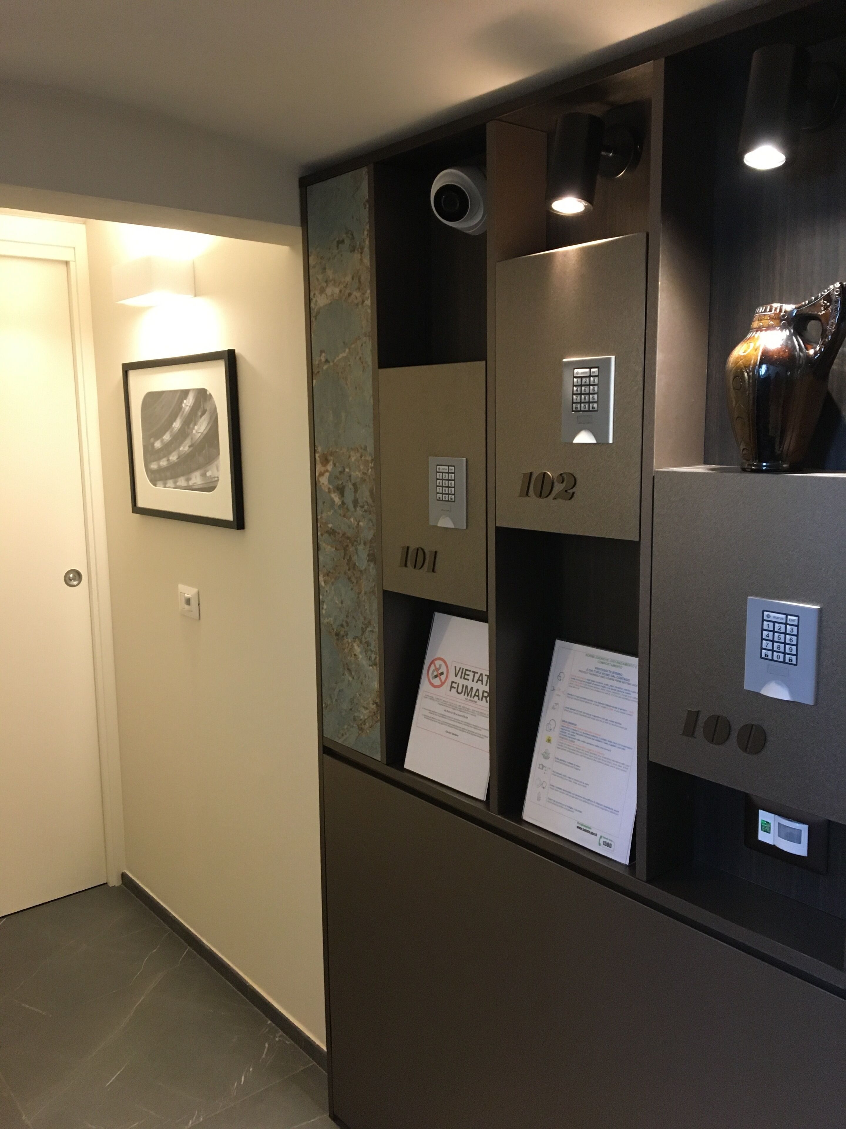 Un couloir orné d’un tableau encadré au mur, un ascenseur dont les portes sont numérotées, et une caméra de surveillance.