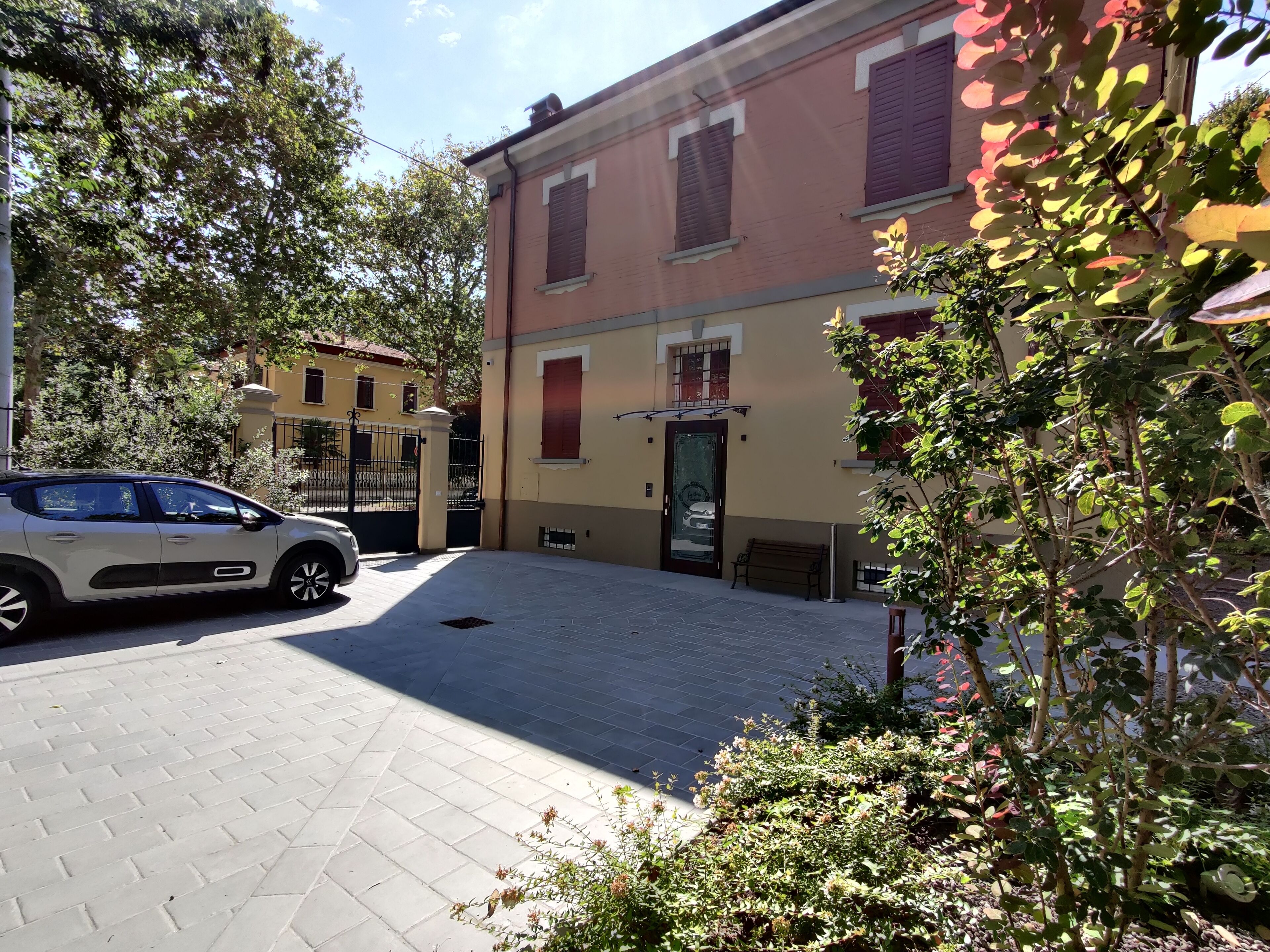 Un patio empedrado con un coche aparcado, un banco y un edificio con puerta y ventanas.
