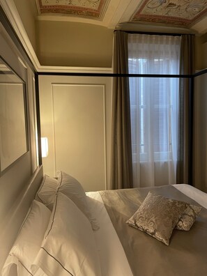 Quarto de hotel com uma cama, uma janela com cortinas translúcidas e teto decorativo.