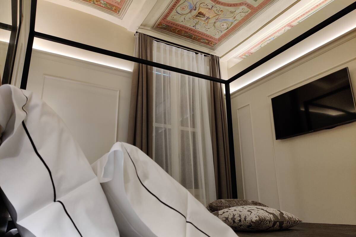 A room with a white sofa, a large window with sheer curtains, a flat-screen TV mounted on the wall, and a decorative ceiling.