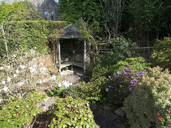 A garden with a wooden gazebo, various shrubs, and flowering plants.