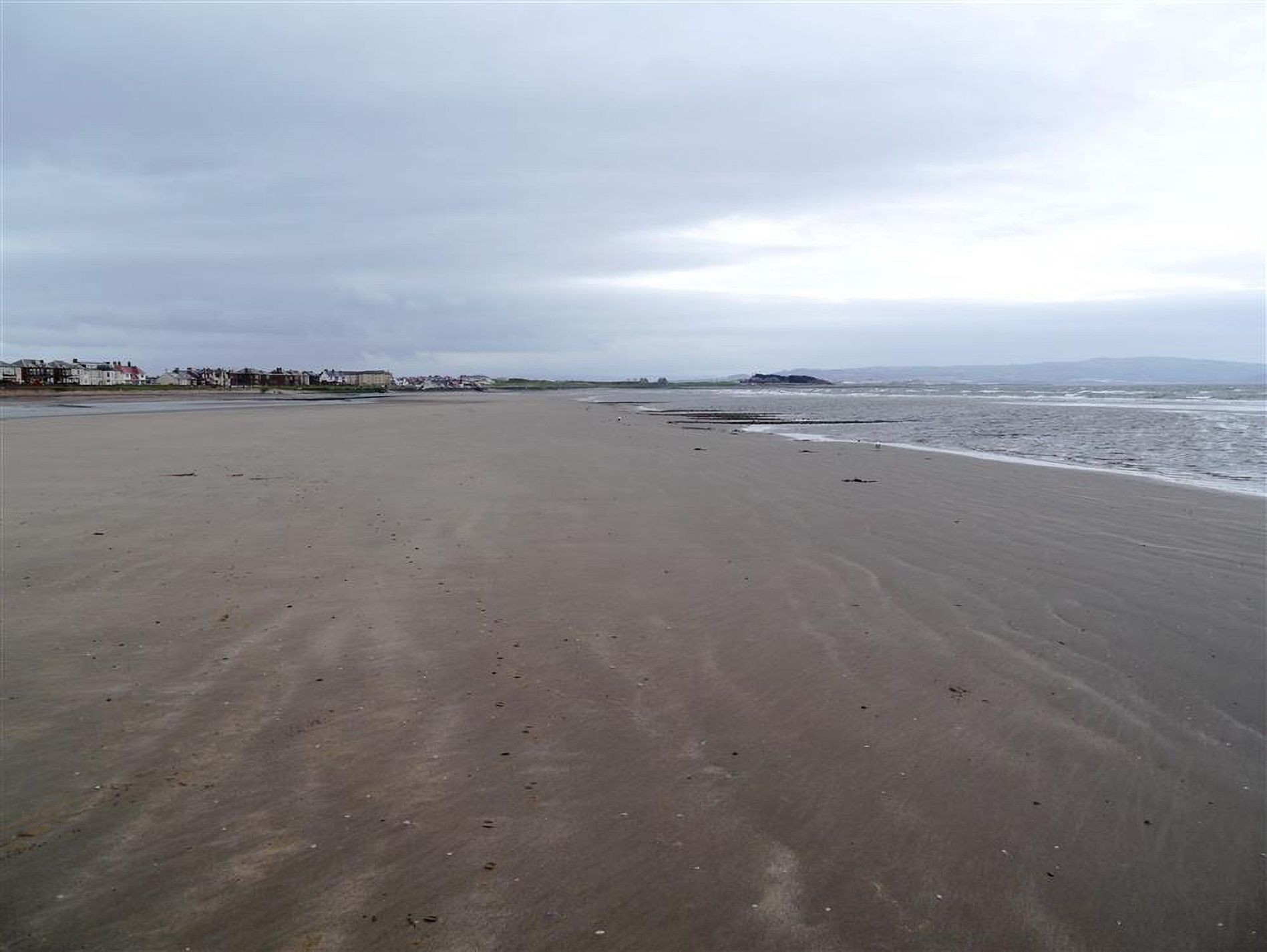 A wide sandy beach with gentle waves, a clear sky, and distant buildings.