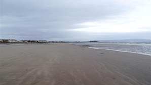 A wide sandy beach with gentle waves, a clear sky, and distant buildings.