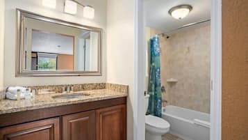 A bathroom with a granite countertop, wooden cabinets, a large mirror, a toilet, and a shower with a blue curtain.