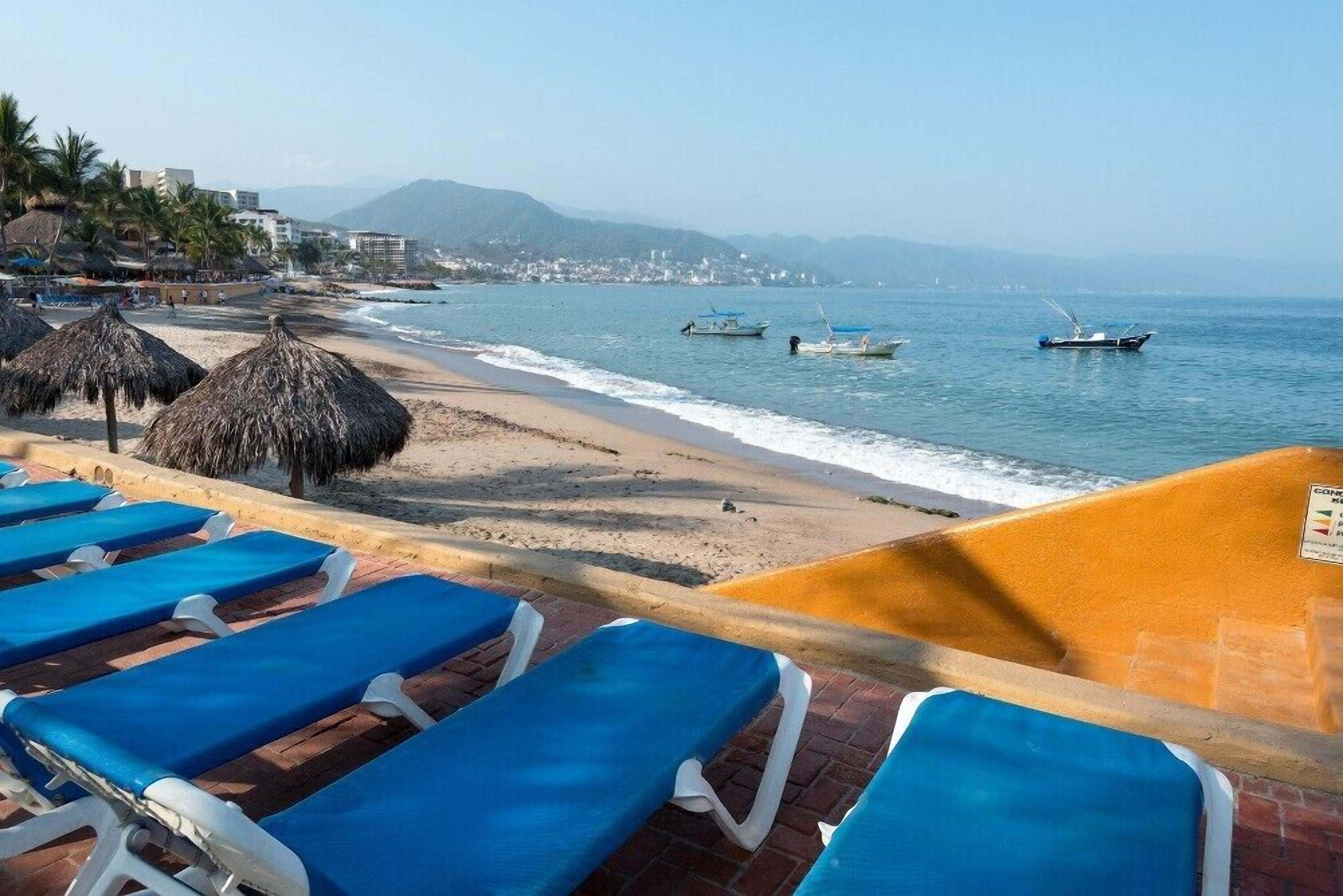 Una playa con sillones de descanso, sombrillas de paja y barcos en el agua.