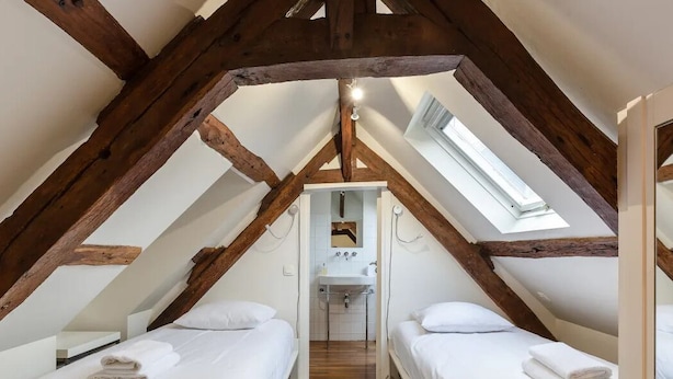 Een slaapkamer met zolderkamergevoel, hoge plafonds, zichtbare houten balken, twee bedden, een dakraam en een kleine badkamer.