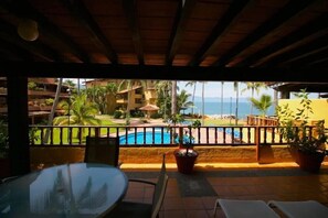 Un balcon donnant sur une piscine et des palmiers, avec des balustrades en bois et des plantes en pot.