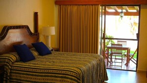 A hotel room with a bed, a nightstand, a lamp, a window with curtains, and a view of an outdoor patio with chairs.