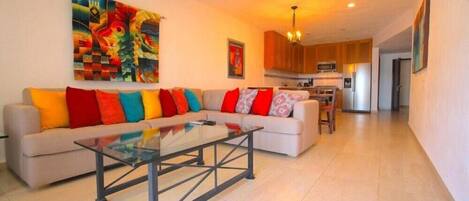 Una sala de estar moderna con un sofá, cojines coloridos, una mesa de centro de vidrio y un cuadro en la pared.