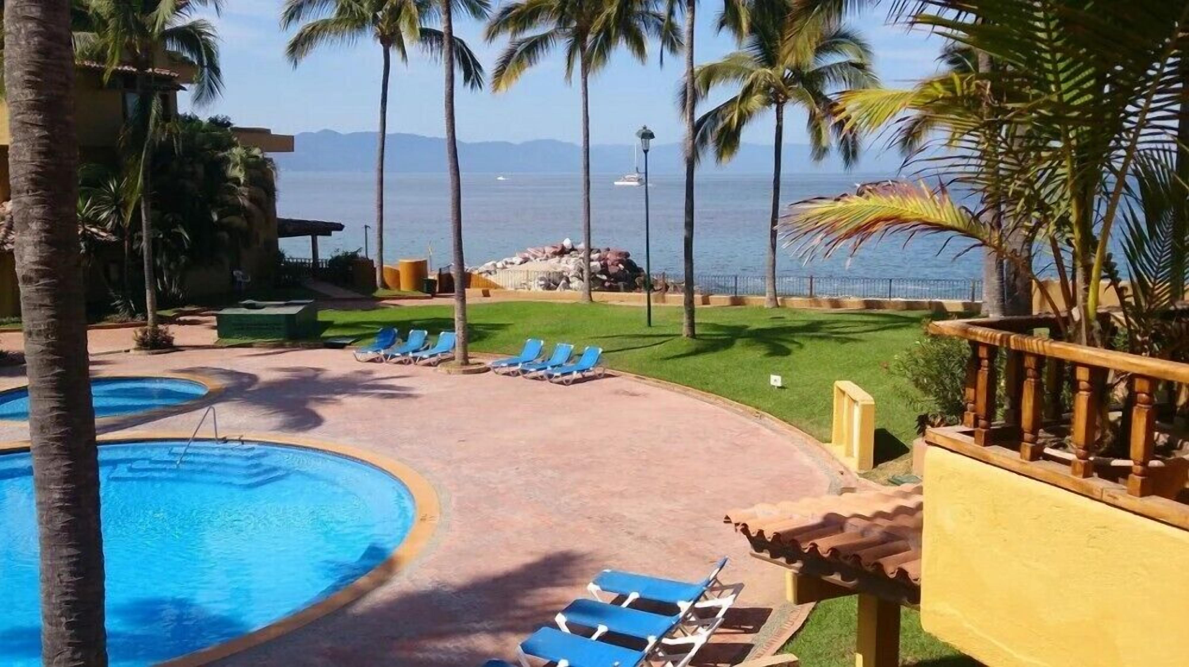 A resort pool area with blue lounge chairs, palm trees, and a view of the ocean.