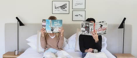 Dos personas leyendo libros sobre una cama con cabecera, cuadros en la pared y mesitas de noche con lámparas.