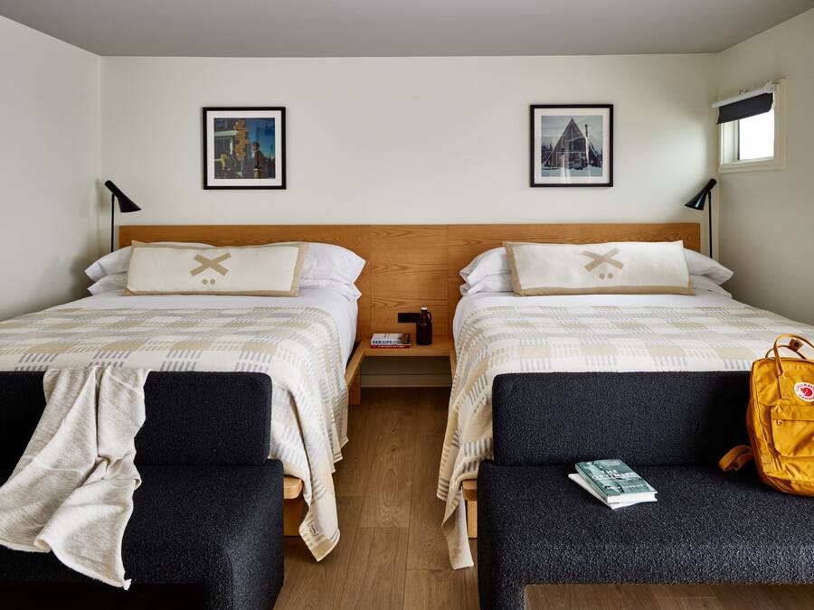 Ein Hotelzimmer mit zwei Betten, einem hölzernen Kopfteil, einem kleinen Nachttisch mit Büchern, einer gelben Tasche und einem Fenster mit Blick.