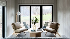 Une pièce chaleureuse avec un plafond haut, des murs en bois et de grandes fenêtres. On y trouve deux fauteuils en cuir, une table ronde ornée d’un vase et de livres, ainsi qu’un lampadaire.