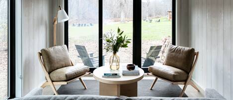 Una habitación acogedora con techo alto, paredes de madera y ventanas amplias. Hay dos sillones de cuero, una mesa redonda con un florero y libros, y una lámpara de pie.