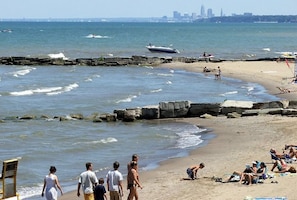 Uma praia com pessoas caminhando e sentadas na areia, um molhe rochoso e barcos na água.