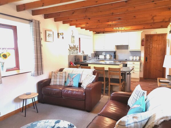 Ein gemütliches Wohnzimmer mit einem braunen Ledersofa, einem kleinen runden Tisch und einem gemusterten Teppich. Im Hintergrund ist eine Küche mit Holzschränken und einer Holztür zu sehen.