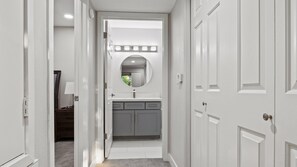 A narrow hallway with a white door on the right, a vanity with a mirror and a sink, and a small window with blinds.