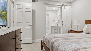 A bedroom with a bed, a dresser, a nightstand, a lamp, and a sliding door leading to a bathroom.