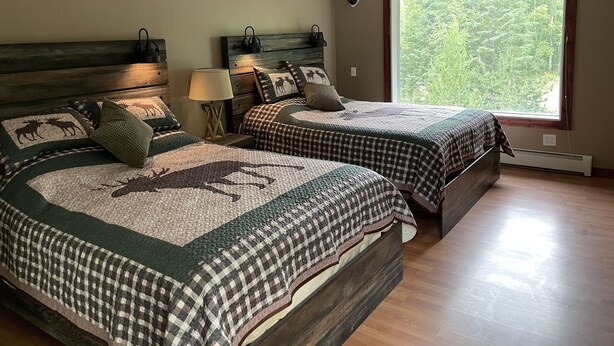 Un dormitorio con dos camas, cada una con colcha estampada de cuadros y diseño de alce, cabeceros de madera, lámparas de noche y un ventanal con vistas a la vegetación.