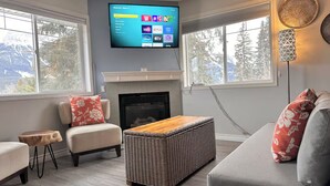 A modern living room with a fireplace, a flat-screen TV mounted above it, and a large window offering a view of trees and mountains.