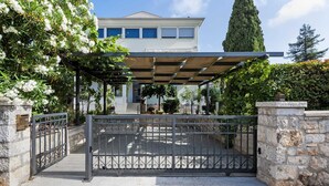 Une entrée fermée par un portail, agrémentée d’une pergola, de murs en pierre et d’un bâtiment visible au loin.