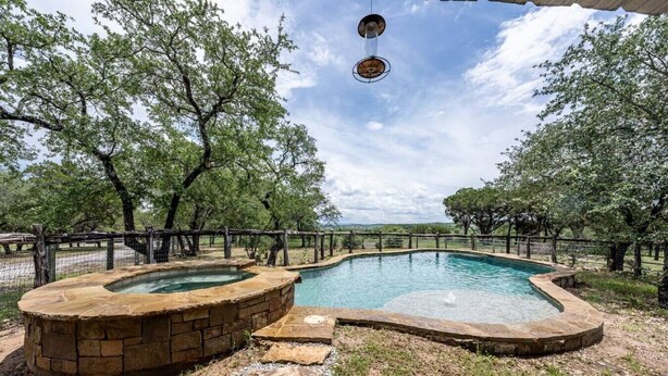 A hot tub with a stone surround, a pool, and a fenced area with trees. - Getaway Ranch - Views for Days +  Pool + Game Room (Spicewood)