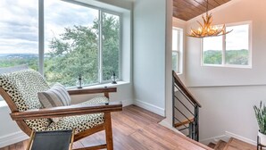 A spacious room with a large window offering a view of the outdoors, a wooden staircase, and a chandelier. - Getaway Ranch - Views for Days +  Pool + Game Room (Spicewood)