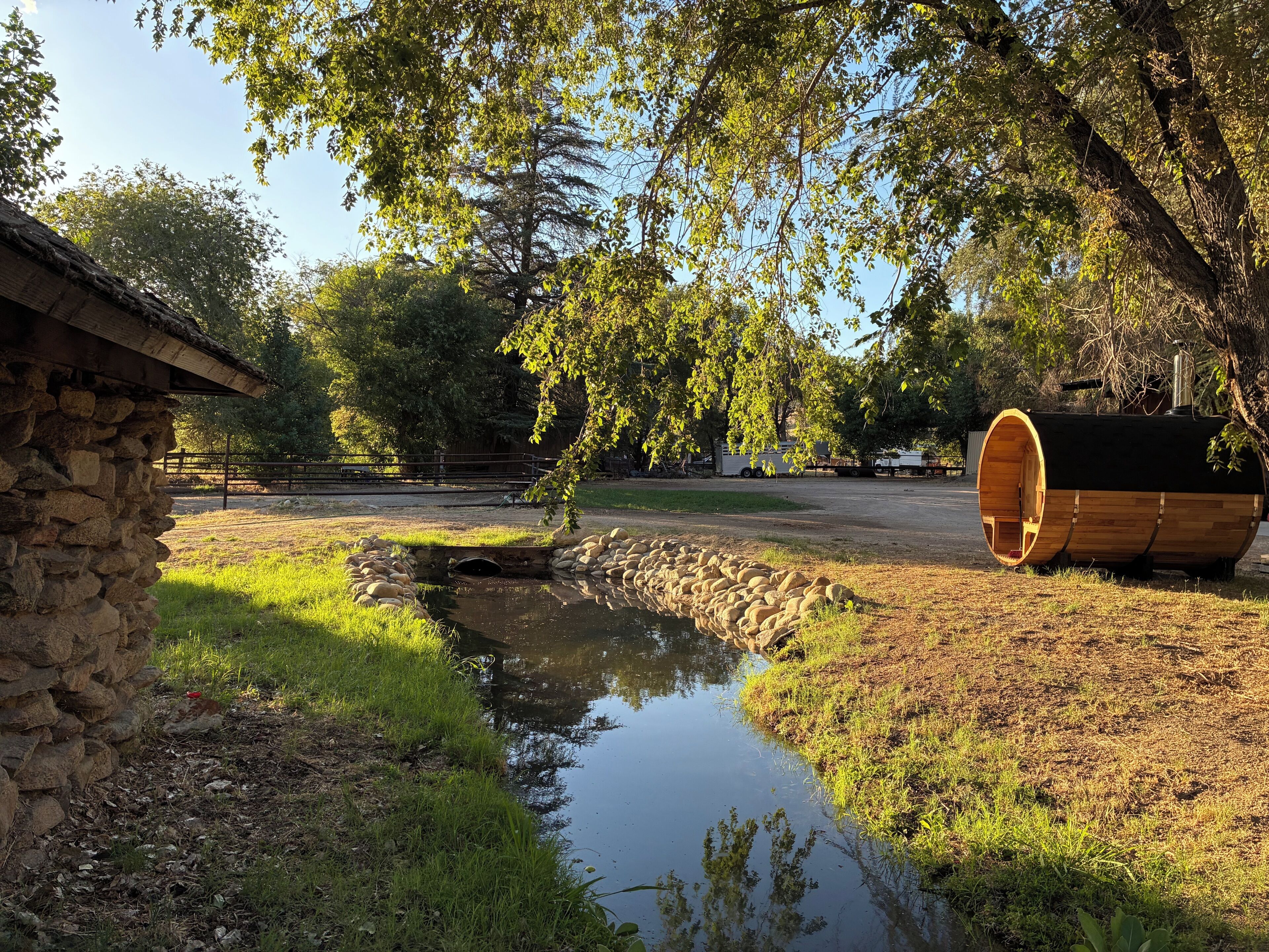 A rustic cabin, a wooden barrel structure, and a small stream in a wooded area.