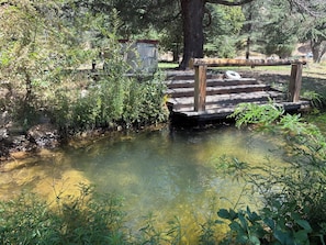 A wooden bridge over a clear stream with surrounding greenery and a small wooden structure.