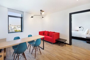Una habitación moderna con suelo de madera, un sofá rojo, un sofá blanco, una mesa de comedor con sillas y vistas a la ciudad a través de una ventana.