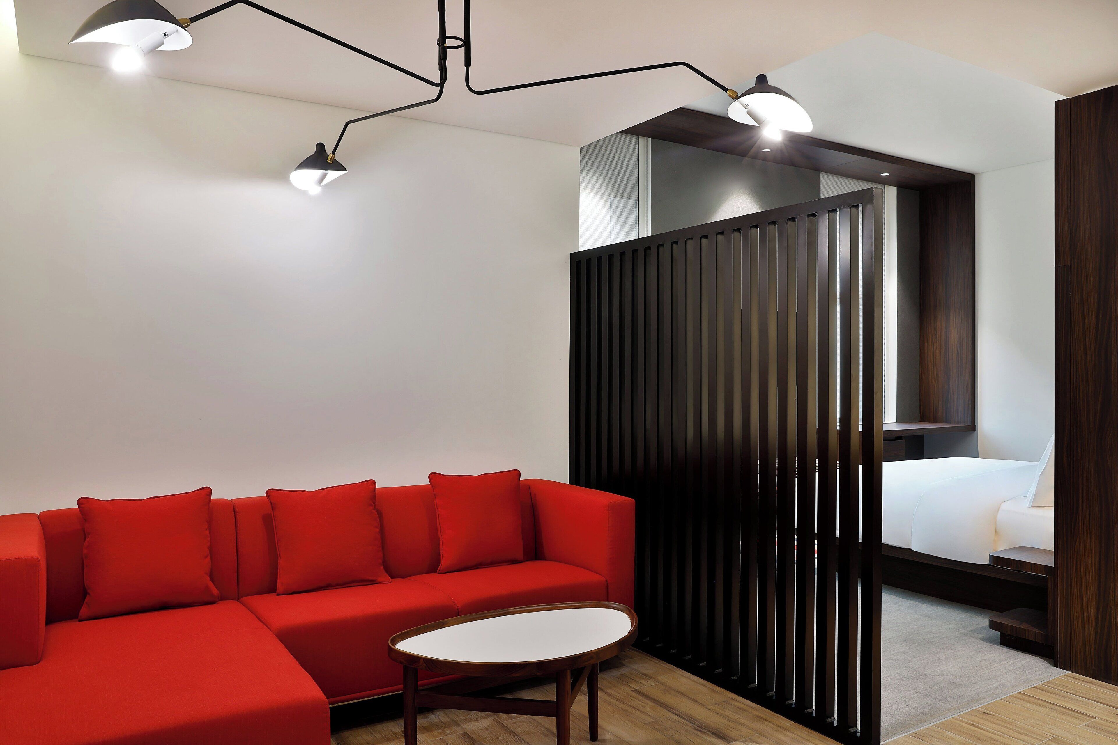 A modern hotel room with a red sectional sofa, a small round table, and a bed with a wooden headboard.