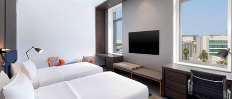 Una habitación de hotel moderna con dos camas blancas, un escritorio, una silla, un televisor y un amplio ventanal con vistas a la ciudad.