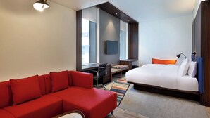 A modern hotel room with a red sofa, a bed with white bedding, a desk with a chair, and a TV mounted on the wall. - Aloft by Marriott Dubai Airport (Dubai)