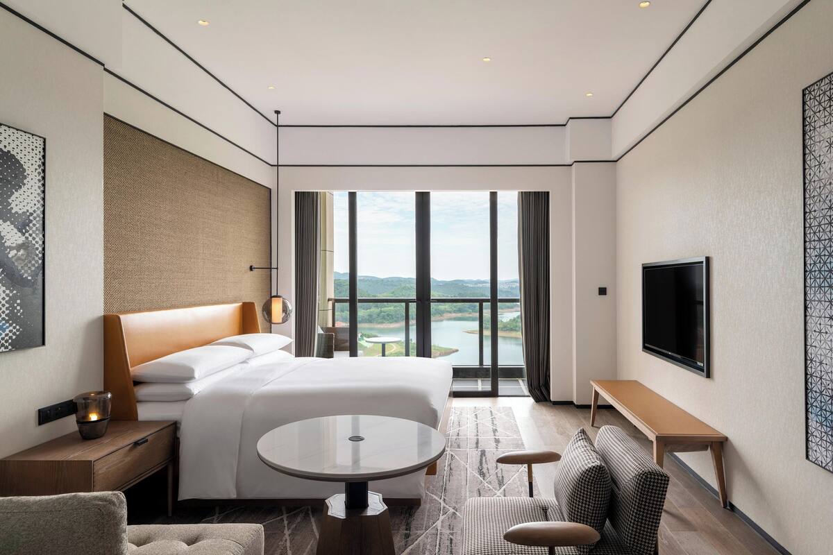 A modern hotel room with a large bed, a TV, a dining table, and a view of a lake.