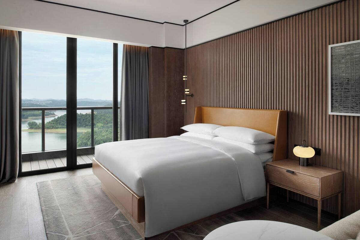 A modern hotel room with a large bed, a nightstand with a lamp, and a view of a lake through floor-to-ceiling windows.
