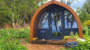 A small, dark blue, arched wooden structure with a door, surrounded by lush greenery and a brick pathway. - Eco Indigo (Dunbar)
