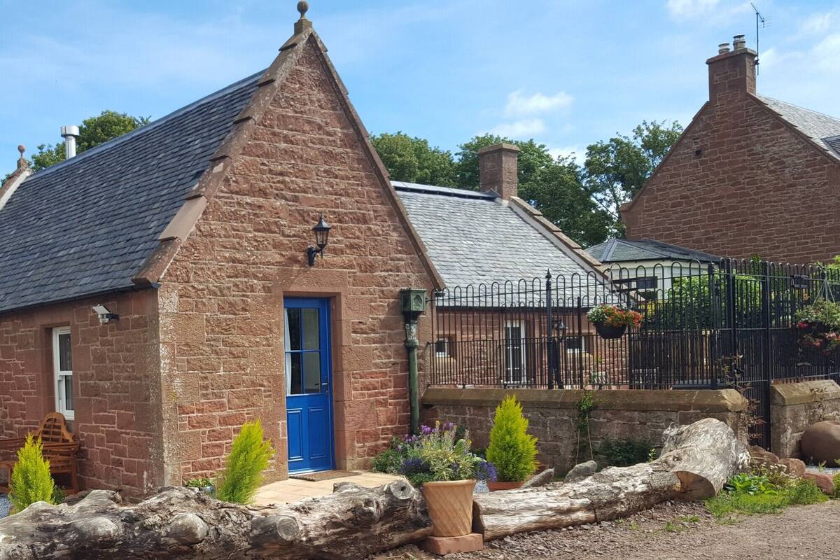 A stone cottage with a blue door, surrounded by a black metal fence, and a garden with various plants and a wooden bench.