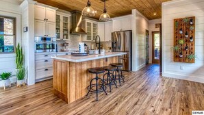 A modern kitchen with wooden flooring, white cabinets, a central island with stools, stainless steel appliances, and pendant lighting.