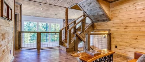 A spacious room with wooden floors, a staircase, and a large window offering a view of greenery.