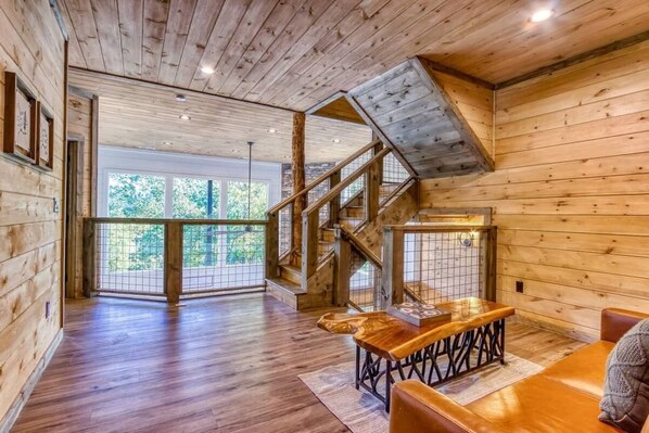 A spacious room with wooden floors, a staircase, and a large window offering a view of greenery.