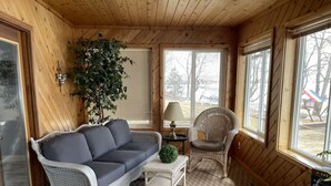 Ein gemütliches Zimmer mit grauem Sofa, einem Korbsessel, einem kleinen Tisch mit Lampe und einem großen Fenster mit Jalousien.