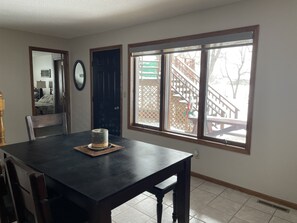 Un coin repas avec une table en bois foncé, une chaise et une fenêtre donnant sur une terrasse en bois et un bâtiment.