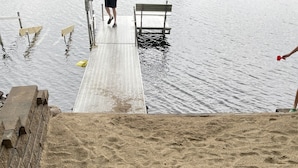 Eine Holzstege führt zu einem Sandstrand mit Stufen und einer Bank.