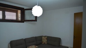 A living room with a sectional sofa, a small table, a ceiling light fixture, a window with blinds, and a wooden door. - Casa vacanze Villa Rosy (nardò)