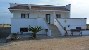 A two-story white building with a tiled roof, a staircase leading to the entrance, and a patio area with a bench and potted plants. - Casa vacanze Villa Rosy (nardò)