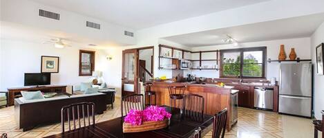 A spacious living area with a dining table, chairs, and a TV. There is a kitchen with modern appliances and a large window.